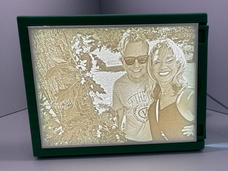 A lithophane we printed, lit from behind in a green frame, showing a smiling couple next to a leafy tree